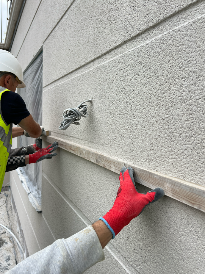 A professional UK plasterer applying a base-coat of through-coloured monocouche render directly to standard brickwork masonry substrate